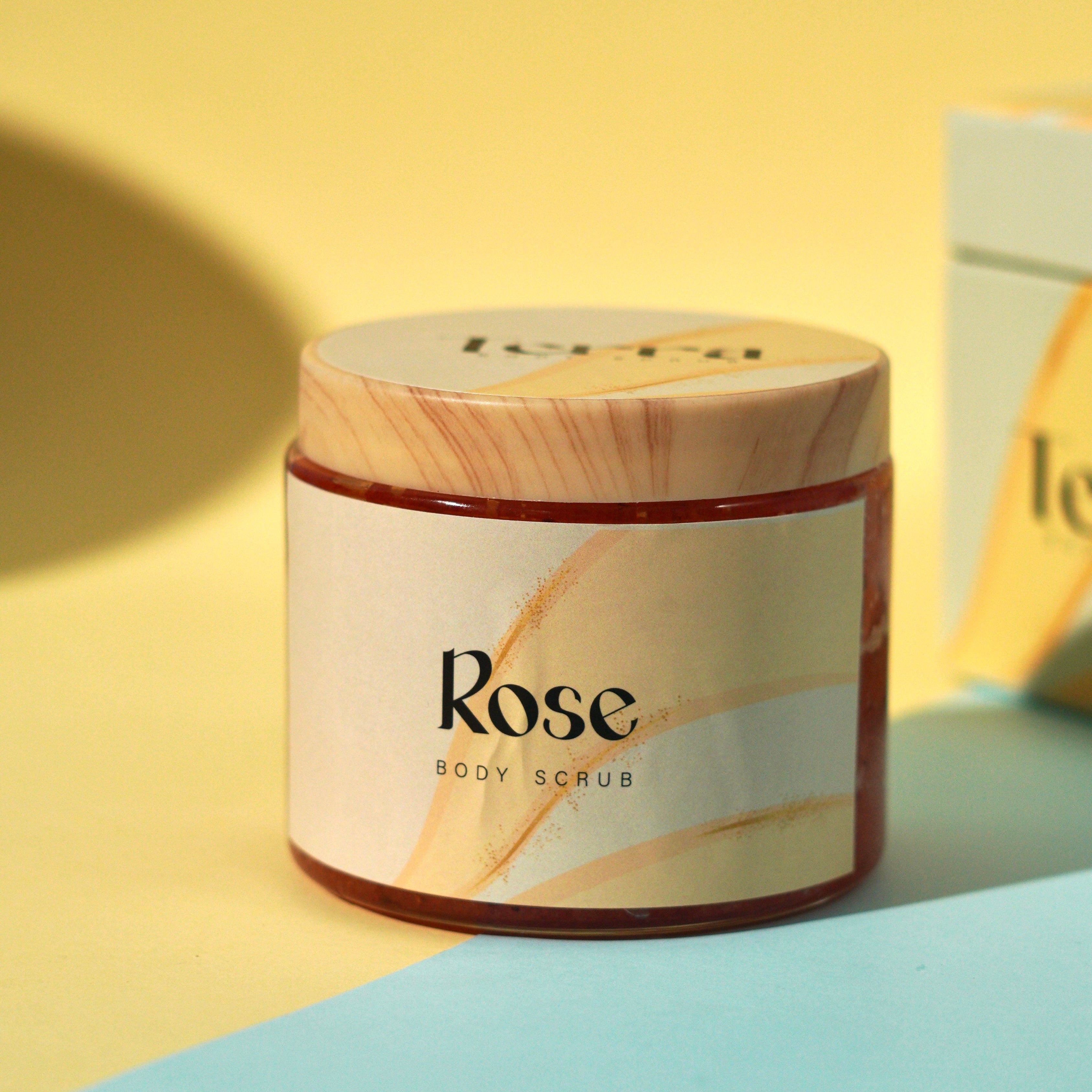 Jar of 'Rose' body scrub with wooden lid on a yellow and blue background